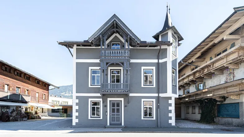 Historisches Gebäude mit grauer Fassade, weißem Putz und einem charmanten Holzbalkon im alpinen Stil.