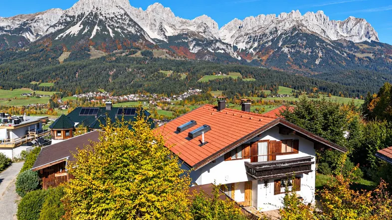 Haus mit rotem Ziegeldach und Balkon, umgeben von Herbstbäumen, mit majestätischem Bergblick.