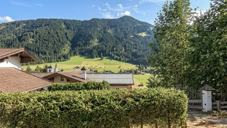 Panoramablick auf die umliegende Berglandschaft mit grünen Wiesen, Wäldern und traditionellen Häusern unter blauem Himmel.