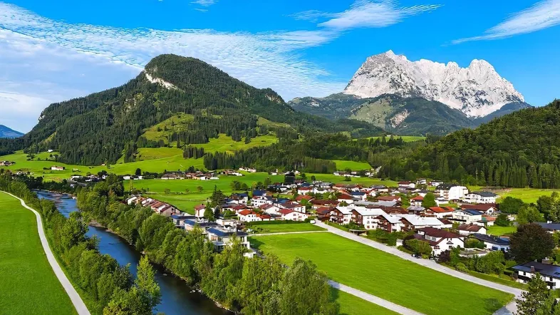 Panoramablick auf ein malerisches Dorf in den Alpen mit Fluss und majestätischen Bergen.