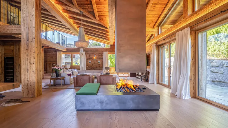 Großzügiges Wohnzimmer im Chalet-Stil mit offenem Kamin, Holzbalken und Zugang zur Terrasse.