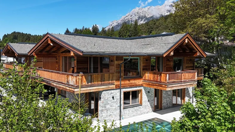 Luxuriöses Holzchalet mit Steinfassade, Balkonen und einem Pool vor alpiner Bergkulisse.