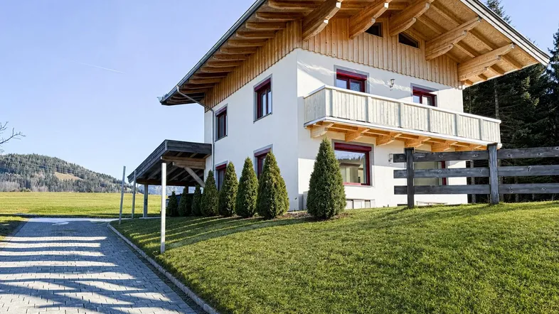 Modernes Haus im alpinen Stil mit weißer Fassade, Holzbalkon und Carport, umgeben von grüner Wiese und Bergen.