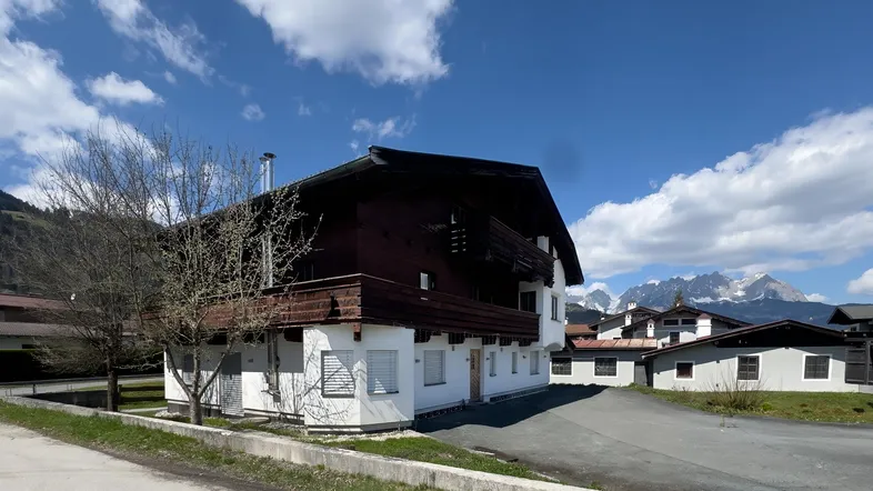 Traditionelles Haus im alpinen Stil mit Holzbalkonen und Blick auf die schneebedeckten Berge unter blauem Himmel.