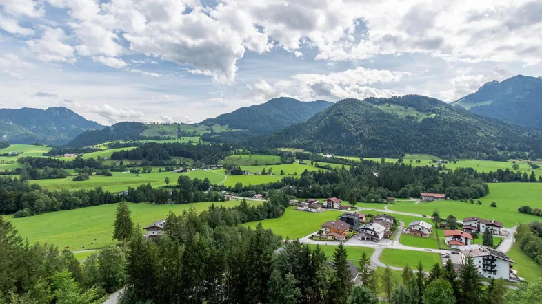 Weitläufige Landschaftsaufnahme mit grünen Wiesen, Wäldern und einem kleinen Dorf unter bewölktem Himmel.