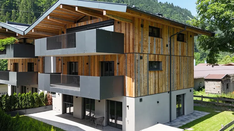 Modernes Mehrfamilienhaus mit Holzfassade, Balkonen und Bergblick in grüner Umgebung.