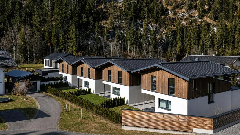 Moderne Reihenhäuser mit Holz- und Weißfassaden, umgeben von grünen Hecken und einem Wald im Hintergrund.