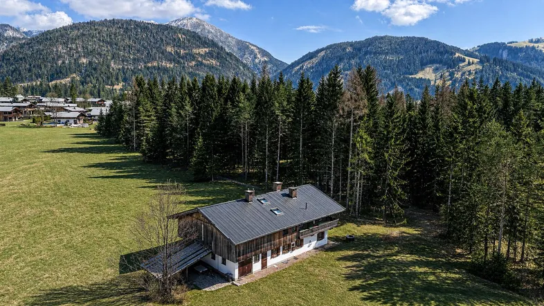 Alpenländisches Haus in grüner Landschaft mit Bergen und Wald im Hintergrund unter blauem Himmel.