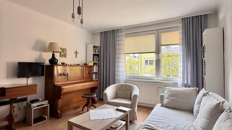 Wohnzimmer mit beigem Sofa, Klavier und Couchtisch, durch große Fenster gut beleuchtet.