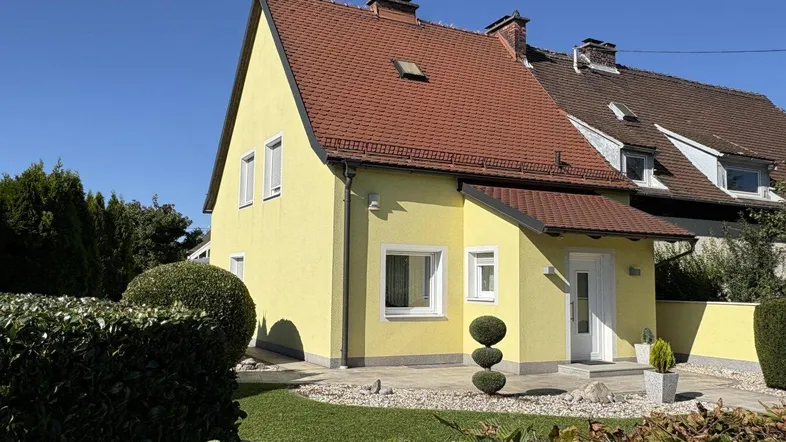 Gelbes Einfamilienhaus mit rotem Ziegeldach und gepflegtem Vorgarten unter blauem Himmel.