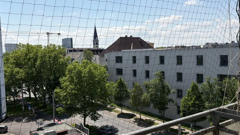 Blick vom Balkon über die Stadt mit Bäumen und Gebäuden im Hintergrund, durch ein Schutznetz.
