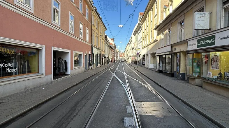 Blick auf eine belebte Stadtstraße mit Straßenbahnschienen und traditionellen Gebäuden unter blauem Himmel.