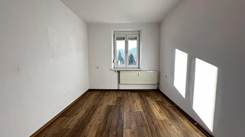 Helles, leeres Zimmer mit Laminatboden und Fensterblick ins Grüne.