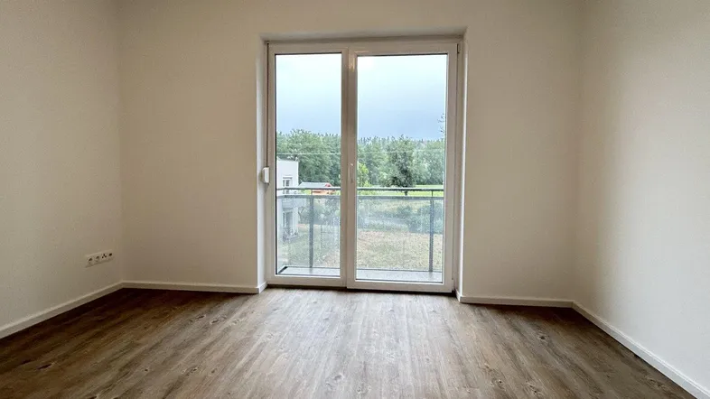 Helles Zimmer mit Laminatboden und Zugang zu einem kleinen Balkon mit Blick ins Grüne.