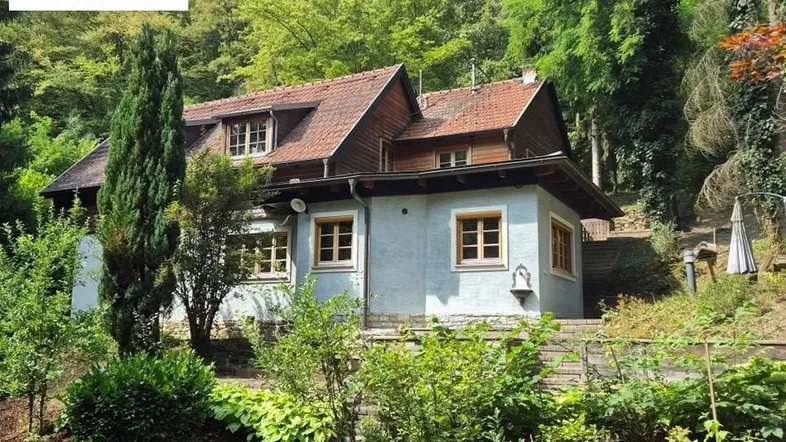 Charmantes Haus mit hellblauer Fassade und rotem Ziegeldach, umgeben von üppigem Grün.