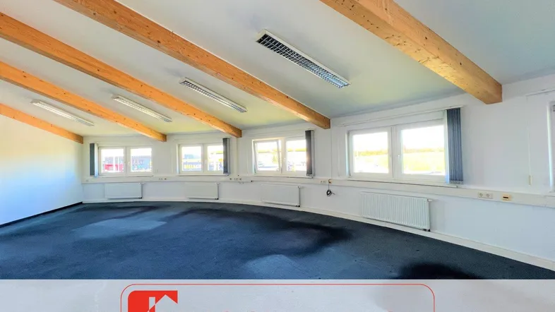 Geräumiger Büroraum mit sichtbaren Holzdeckenbalken, vielen Fenstern, Teppichboden und Blick ins Grüne.