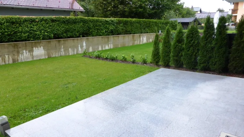 Große gepflasterte Terrasse mit angrenzendem Rasenbereich und einer hohen Hecke im Hintergrund.