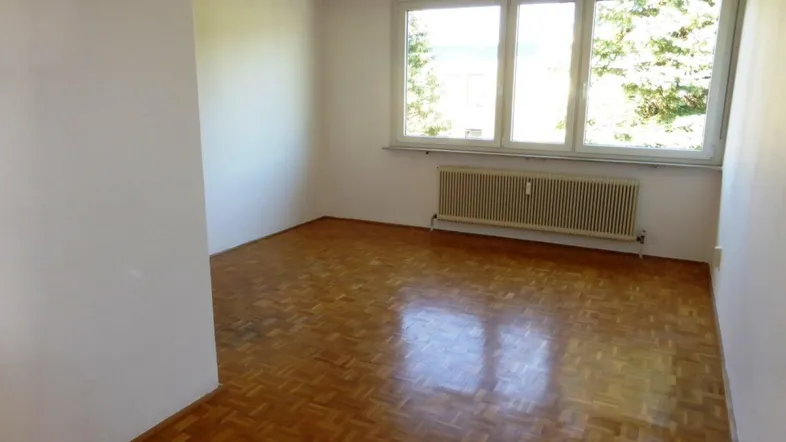 Helles Zimmer mit Parkettboden, weißen Wänden und großem Fenster mit Blick ins Grüne.
