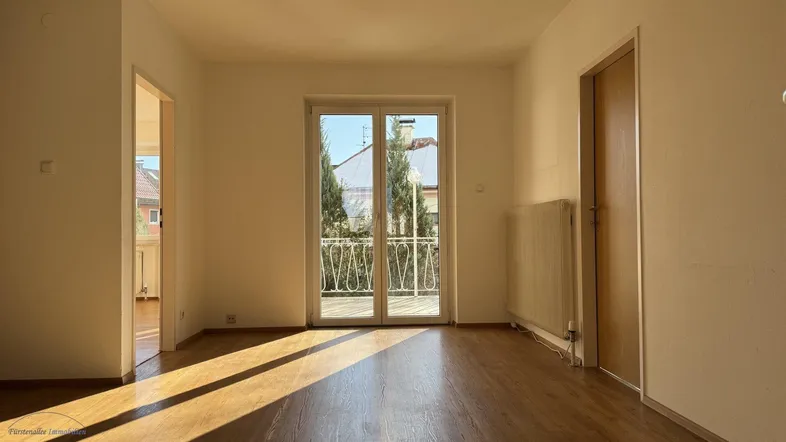 Helles Wohnzimmer mit Holzboden und Zugang zu einem kleinen Balkon, durchflutet von Sonnenlicht.
