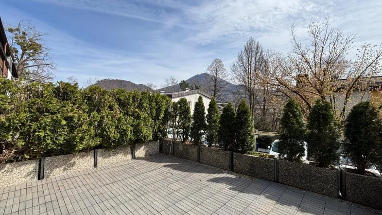 Große gepflasterte Terrasse mit Hecke und Blick auf die umliegende Berglandschaft unter blauem Himmel.