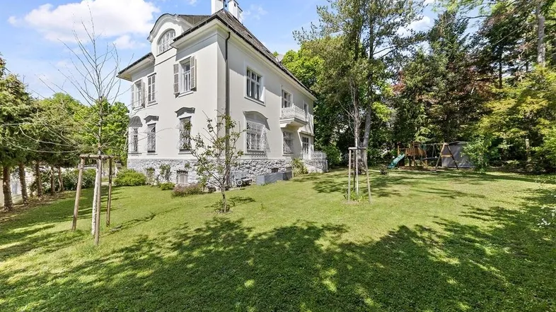 Weißes Herrenhaus mit großem Garten und Spielplatz unter blauem Himmel.