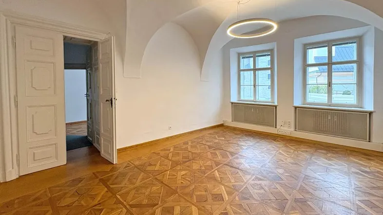 Geräumiges Zimmer mit Gewölbedecke, Fischgrätparkett und zwei großen Fenstern, das viel Tageslicht hereinlässt.