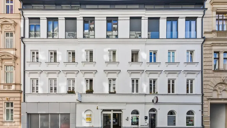 Weißes Mehrfamilienhaus mit moderner Architektur, vielen Fenstern und Rollläden an der Fassade.