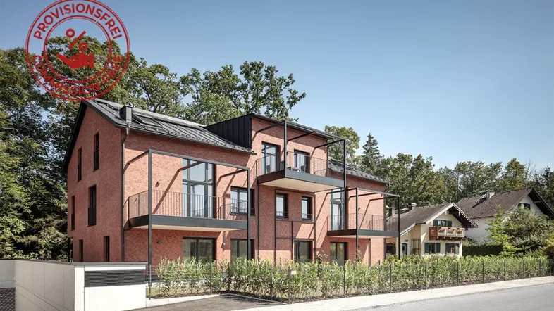 Modernes Mehrfamilienhaus mit roter Ziegelfassade und mehreren Balkonen in grüner Umgebung.