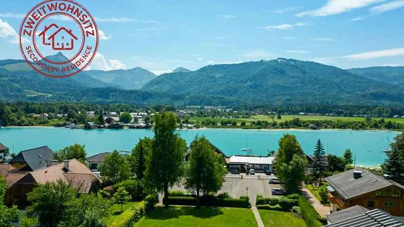 Panoramablick auf den See und die umliegende Berglandschaft mit Häusern und Grünflächen.