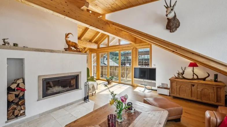 Gemütliches Wohnzimmer im Dachgeschoss mit Holzbalken, Kamin und Zugang zum Balkon.