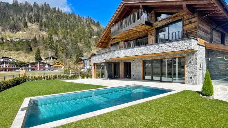 Modernes Chalet mit Pool und Garten, umgeben von Bergen unter blauem Himmel.
