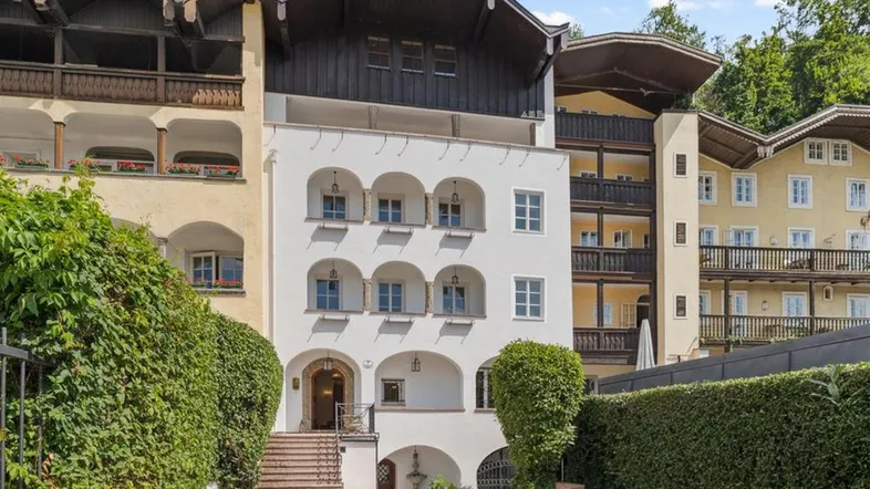 Traditionelles Gebäude mit weißer Fassade, Holzbalkonen und gepflegter Hecke am Eingang.