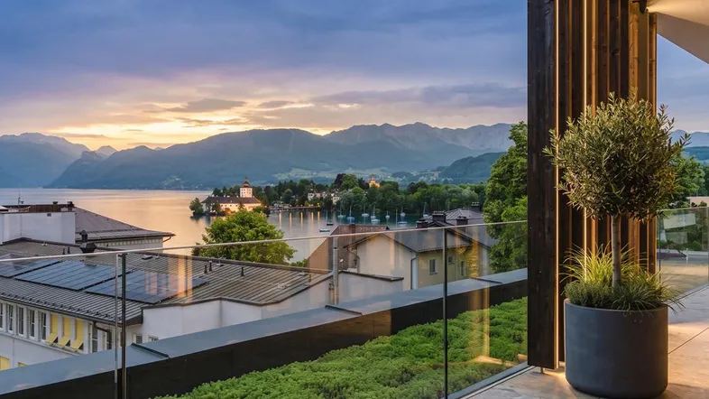 Moderner Balkon mit Glasgeländer, Pflanzen und atemberaubendem Blick auf den See und die Berge bei Sonnenuntergang.