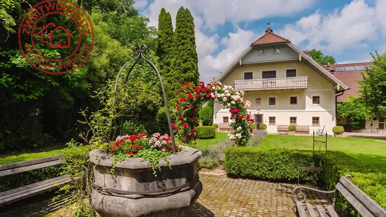 Traditionelles Haus mit gepflegtem Garten, Zierbrunnen und Rosenbogen unter blauem Himmel.
