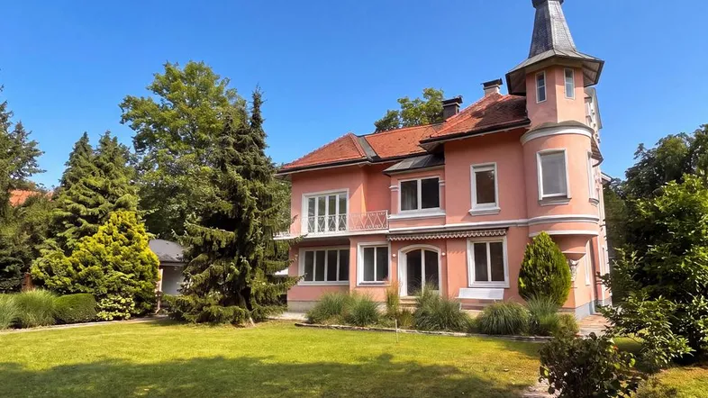 Elegante rosa Villa mit markantem Turm und gepflegtem Vorgarten unter blauem Himmel.
