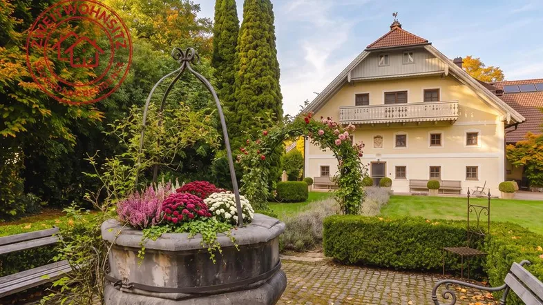 Malerischer Garten mit altem Brunnen und blühenden Pflanzen vor einem traditionellen Landhaus.