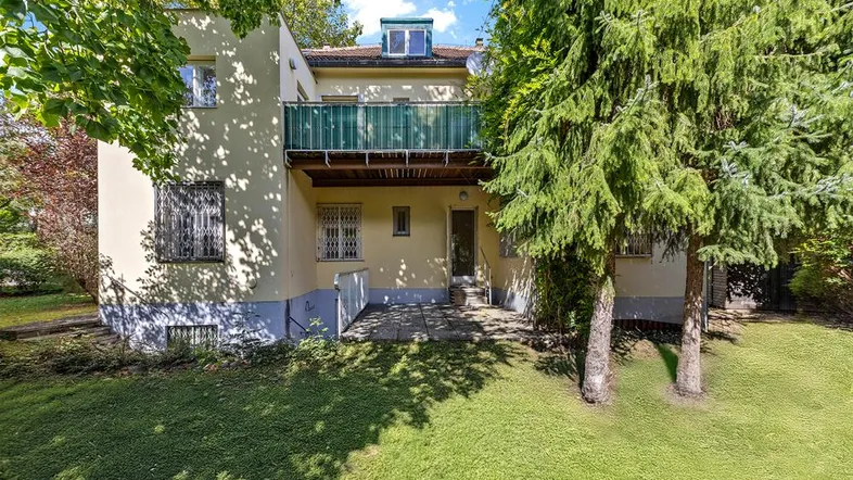 Hinteransicht eines gepflegten Hauses mit grünem Garten, gepflasterter Terrasse und einem Balkon.