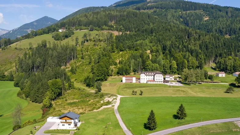 Luftaufnahme eines weitläufigen Anwesens mit mehreren Gebäuden, umgeben von grünen Wiesen und bewaldeten Bergen.