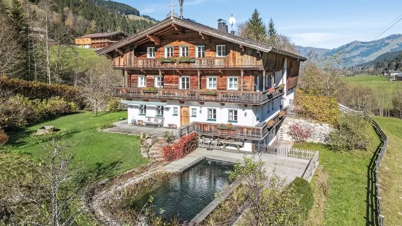 Traditionelles Holzhaus mit Naturpool und Bergblick, eingebettet in eine grüne Hügellandschaft unter blauem Himmel.