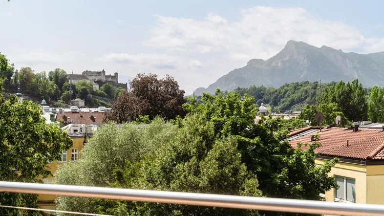 Blick von der Terrasse über die Stadt Salzburg mit Festung Hohensalzburg und umliegenden Bergen.