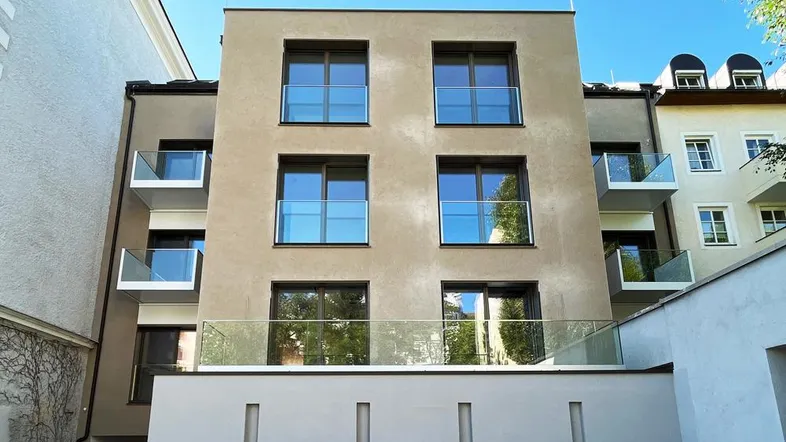 Moderne Gebäudefassade mit großen Fenstern, Glasbalkonen und einer Terrasse im Erdgeschoss.