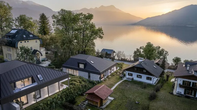 Luftaufnahme von Häusern am Seeufer mit Bergen im Hintergrund bei Sonnenuntergang.