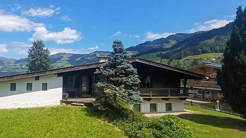 Traditionelles Haus im alpinen Stil mit Bergblick und grüner Umgebung unter blauem Himmel.