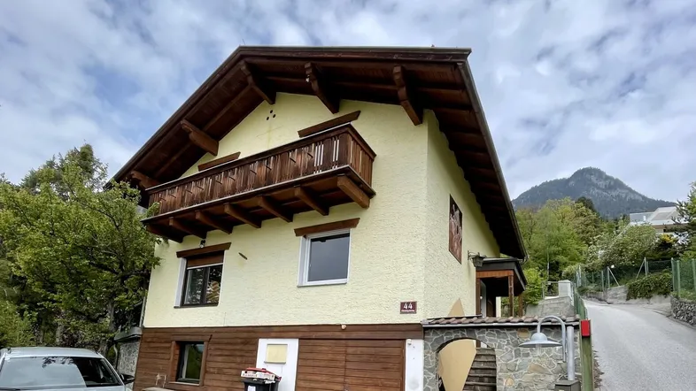 Traditionelles Haus mit Holzbalkonen und Satteldach, umgeben von grüner Landschaft und Bergen.