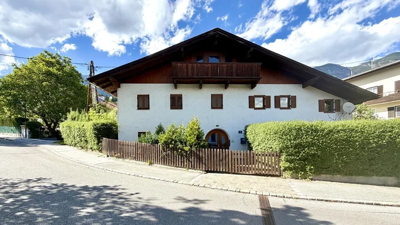 Traditionelles Haus mit weißer Fassade, Holzbalkon und gepflegter Hecke unter blauem Himmel.