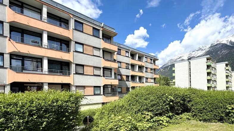 Mehrfamilienhaus mit Balkonen und Blick auf die schneebedeckten Berge unter blauem Himmel.