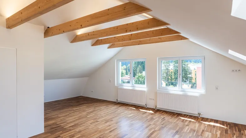 Helles Dachgeschosszimmer mit Holzbalken, Parkettboden und Fenstern mit Blick ins Grüne.