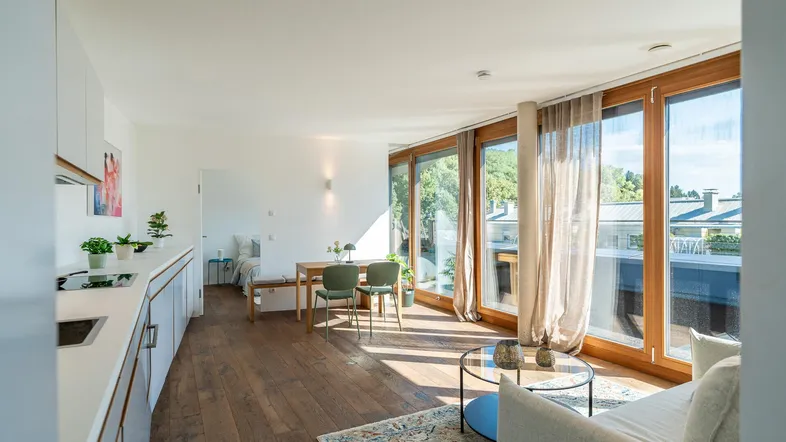 Helles, offenes Wohn-Esszimmer mit integrierter Küche, Holzboden und großen Fenstern mit Blick ins Grüne.