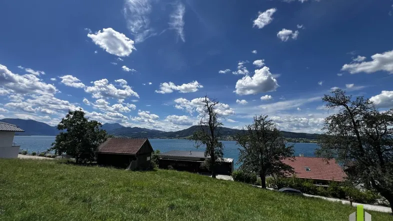 Panoramablick über einen großen See mit umliegenden Bergen und grünen Wiesen unter blauem Himmel.