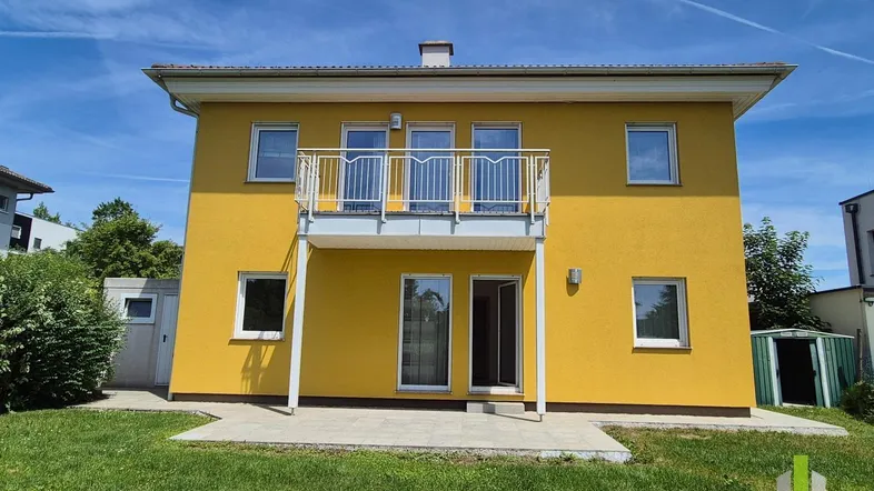 Gelbes Einfamilienhaus mit Balkon, kleiner Terrasse und grünem Garten unter blauem Himmel.